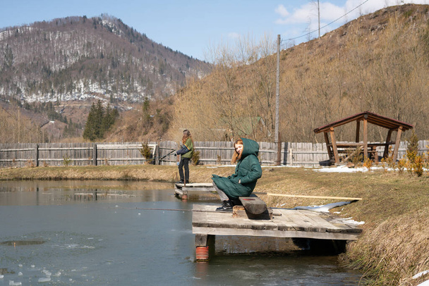Семейная рыбалка Молодые люди сидят с удочками на деревянных пирсах у озера, покрытого тонким льдом. Рыбалка форели. Карпаты. Украина. - Фото, изображение