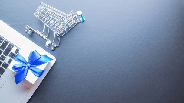 Online nákupní byt. Notebook, nákupní vozík a bílý dárek s modrou stuhou na tmavém pozadí. Internetový nákup, koncept internetového obchodu. Internetový obchod - Fotografie, Obrázek