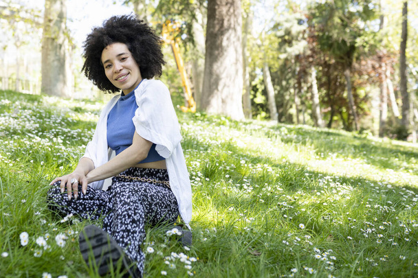 pretty mixed afro woman sitting on the grass, in a park in spring - Photo, Image