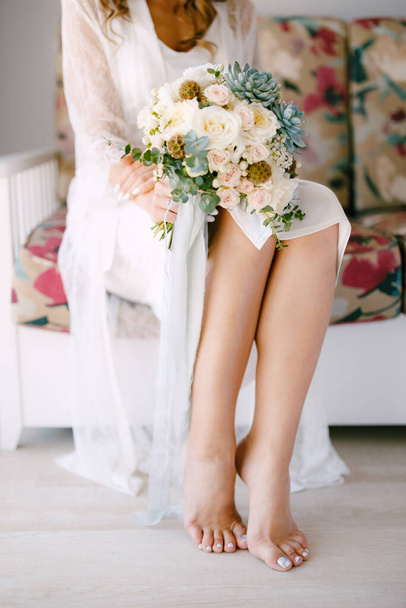 Barefoot bride in a lace robe sits on a sofa in a hotel room and holds a wedding bouquet in her hands, close-up - Photo, Image
