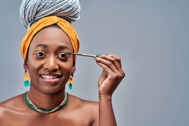 Smiling girl applies black mascara on her eyelashes. Photo of young african girl finishing her makeup on grey background. Beauty concept - Фото, изображение