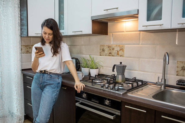 nő iszik reggeli kávé tea a konyhában szörfözés interneten a telefonon - Fotó, kép