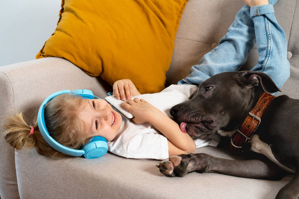 Mała blondynka słucha muzyki ze słuchawkami leżącymi na kanapie z dużym psem i uśmiechnięta, pies liże jej rękę, koncepcja miłości do zwierząt. - Zdjęcie, obraz
