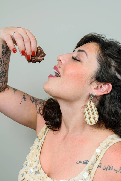 Tetovált nő nyitott szájjal és csukott szemmel, kezében egy brazil mézes tortával a feje felett. Tetoválás a bal vállára írva arabul - Apa, Fiú és Szentlélek. - Fotó, kép