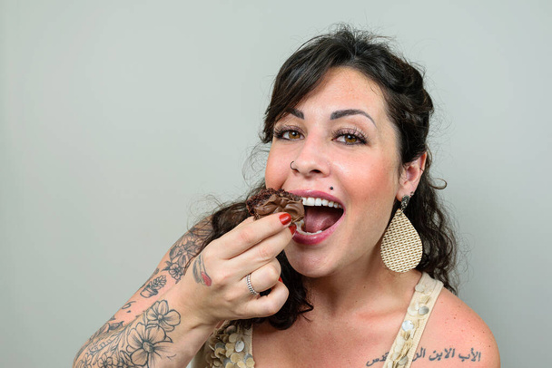 入れ墨、口を開けて笑顔の女性は、ブラジルの蜂蜜のケーキを食べる準備ができている。アラビア語で書かれた左肩の入れ墨 – 父,息子と聖霊. - 写真・画像