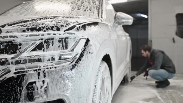 Espuma gotas de un SUV moderno durante el lavado de coches. Hombre usando cepillo para limpiar la superficie de la llanta de la rueda de un coche en el lavado automático - Metraje, vídeo