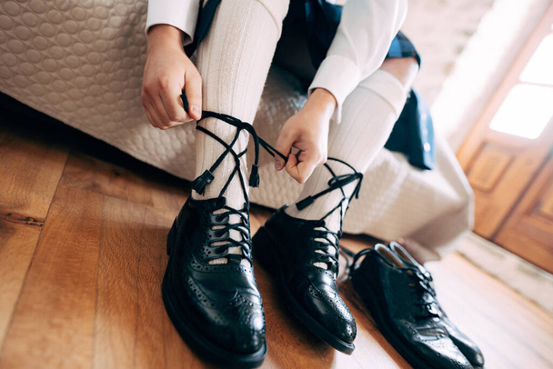 Preparándose para una boda escocesa. El hombre con un kilt y calcetines altos se sienta en la cama atando cordones largos en sus zapatos. Cerca hay un par de zapatos negros - Foto, Imagen