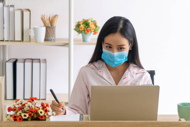 Die freiberufliche junge Frau trägt Masken, die vor Luftkrankheiten und bei Ausbruch des Covid 19 schützen, während sie von zu Hause aus mit Laptop arbeitet. Gesellschaftliche Distanz verhindert die Ausbreitung des Coronavirus. - Foto, Bild
