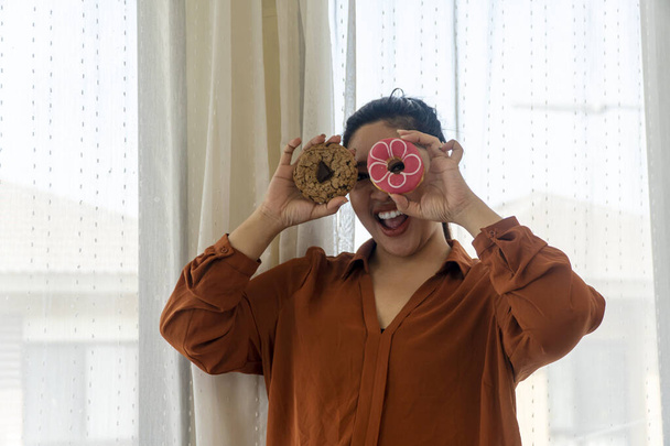 Солодощі шкідливі для здорової концепції. Щаслива усмішка ожиріння товста жінка тримає печиво з шоколадними чіпсами і пончик в руці прикриває власні очі, стоячи в кімнаті вдома
. - Фото, зображення
