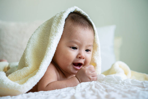 Neşeli şirin bebek beyaz battaniyenin altında kameraya bakıyor. Masum bebek, beyaz yatakta kafasında havluyla sürünüyor.. - Fotoğraf, Görsel