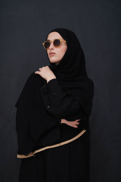 Joven musulmán en ropa tradicional o abaya y gafas de sol posando frente a pizarra negra. Mujer árabe que representa el estilo de vida árabe moderno, la moda islámica y el concepto de kareem Ramadán - Foto, imagen