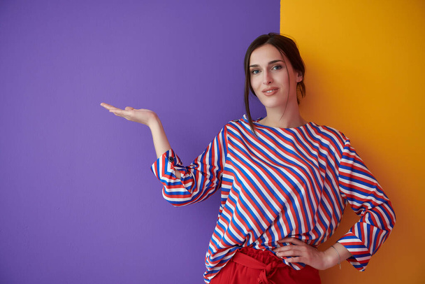 Portrait of happy smiling young beautiful woman in a presenting gesture with open palm isolated on purple and yellow background. Female model in modern fashionable clothes posing in the studio - Photo, Image