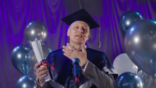педагог-мужчина в академической одежде поздравляет студентов с окончанием учебы, говорит в микрофон, стоя в конференц-зале на фоне воздушных шаров - Кадры, видео