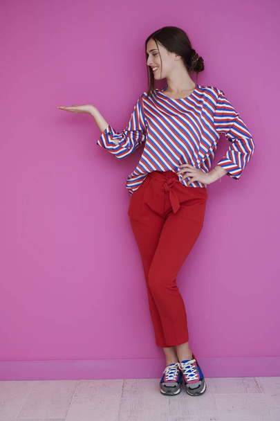 Portrait of happy smiling young beautiful woman in a presenting gesture with open palm isolated on pink background. Female model in modern fashionable clothes posing in the studio - Photo, image