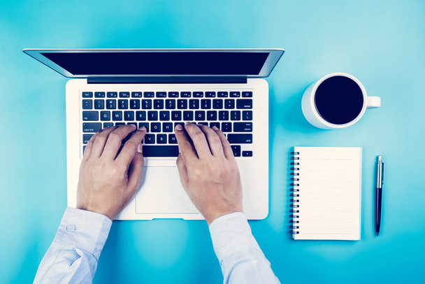 Flat lay, ręka biznesmena pracującego na laptopie z filiżanką kawy na biurku w biurze, miejscu pracy i notebooku oraz książka na niebieskim tle, przestrzeń robocza i przestrzeń do kopiowania, widok z góry, koncepcja biznesowa. - Zdjęcie, obraz