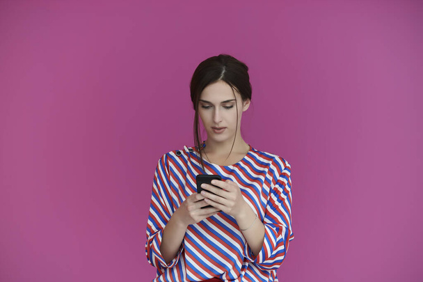 Retrato de una chica joven con cara seria mientras usa un teléfono inteligente aislado sobre un fondo rosa. Mujer modelo de chat o navegación en las redes sociales. Concepto de moda y tecnología - Foto, imagen