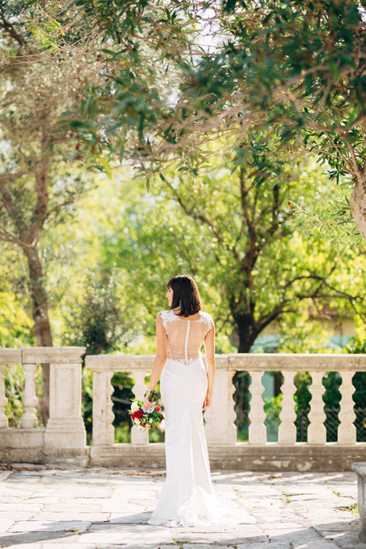 Novia en un vestido blanco con un ramo de flores está de pie con su espalda en el balcón sobre el fondo de árboles verdes - Foto, imagen