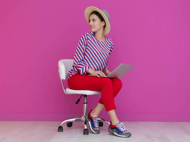 Porträt einer jungen Frau, die auf dem Stuhl sitzt und ihren Laptop auf dem Schoß hält, isoliert auf rosa Hintergrund. Weibliches Model präsentiert Mode und technologisches Konzept - Foto, Bild