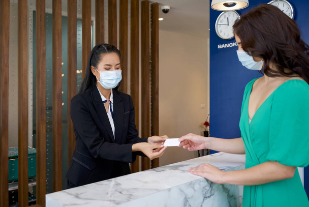 El gerente del hotel en traje negro lleva una máscara mientras le entrega una tarjeta de habitación al huésped. La pared trasera tiene un reloj para decir la hora en la ciudad capital de Tailandia. - Foto, Imagen