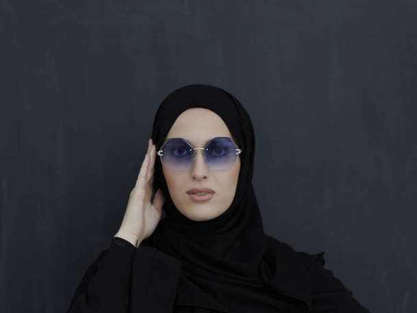 Young muslim in traditional clothes or abaya and sunglasses posing in front of black chalkboard. Arab woman representing modern arabic lifestyle, islamic fashion and Ramadan kareem concept - Foto, imagen