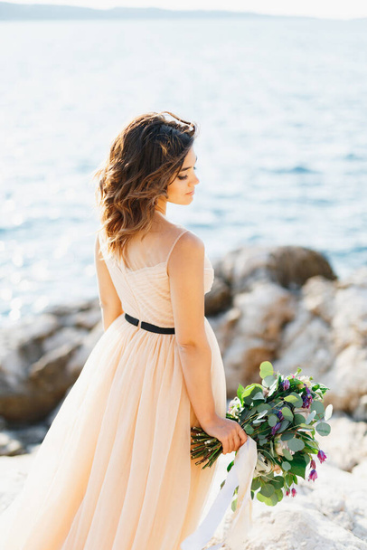 Gyönyörű, töprengő menyasszony áll oldalra egy csokor virággal egy sziklán a tenger felett. - Fotó, kép