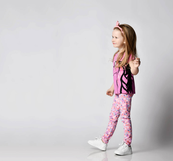 Happy frolic blonde kid girl in summer clothing colorful pants and t-shirt walks looking at copy space. Side view - Fotó, kép