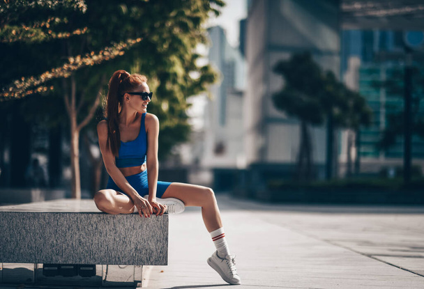 Smiling happy athlete woman in sportswear runner sits on bench in city after workout exercises and looks aside in Dubai - Φωτογραφία, εικόνα