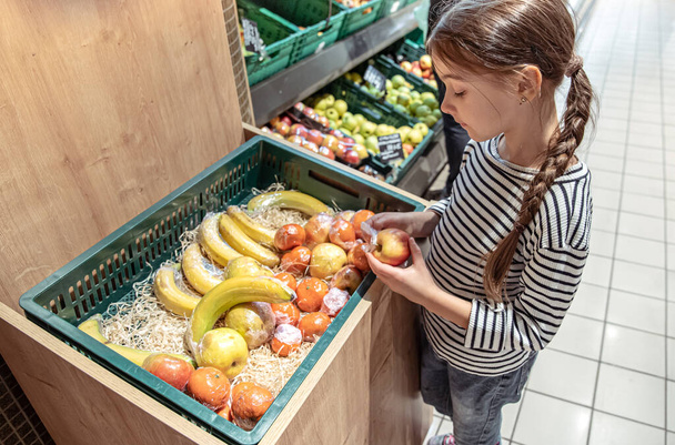 Una niña elige manzanas y plátanos envueltos individualmente en la tienda. - Foto, Imagen