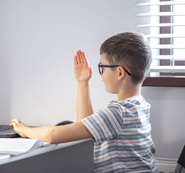 Een basisschoolstudent met een bril zit aan een tafel met een laptop, leert op afstand, steekt zijn hand op in een online les. - Foto, afbeelding