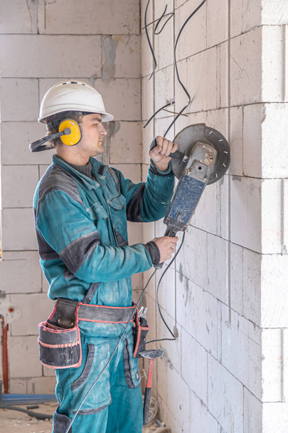 Un operaio edile elettricista in un casco protettivo in una struttura di lavoro lavora con una smerigliatrice. - Foto, immagini