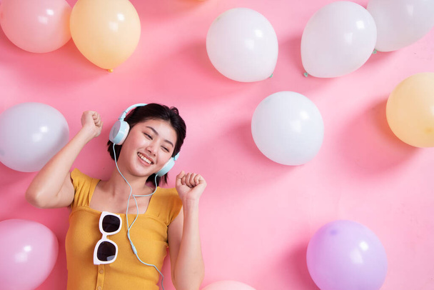 Renkli hava balonlarıyla pembe arka planda gülen Asyalı güzel kız. Doğum günü partisindeki genç kadın. Pastel renkli balonla kutluyor. Müzik dinler ve dans eder.. - Fotoğraf, Görsel