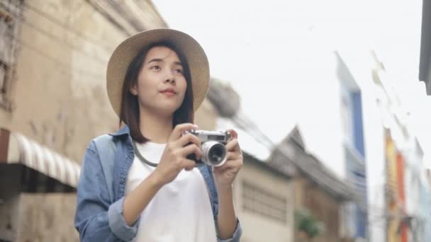 Mujer feliz joven viajero asiático con sombrero retro fedora usando cámara de cine tomando una foto en una pequeña calle en Bangkok Tailandia. Disfrutando viajar en verano de vacaciones. Concepto de viaje individual. - Metraje, vídeo