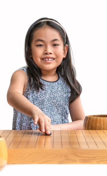 Go oynayan sevimli Asyalı kız çocuğu portresi ya da Japon satranç tahtası oyunu, ya da Çin satranç tahtası oyunu, kameraya bakıp gülümseme, beyaz arka plan, dikey görüntü. - Fotoğraf, Görsel
