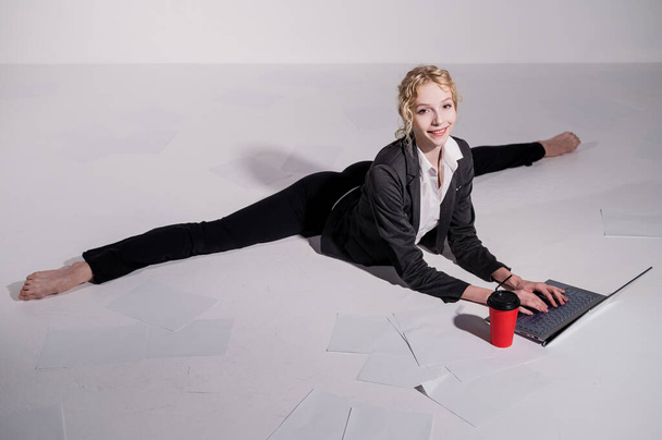 A ballerina dressed in a business suit sits in a middle twine, works at a laptop and drinks coffee. Flexible woman works at the computer - Foto, Imagen
