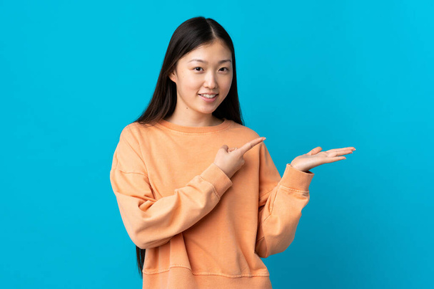 Young Chinese girl over isolated blue background holding copyspace imaginary on the palm to insert an ad - Foto, afbeelding