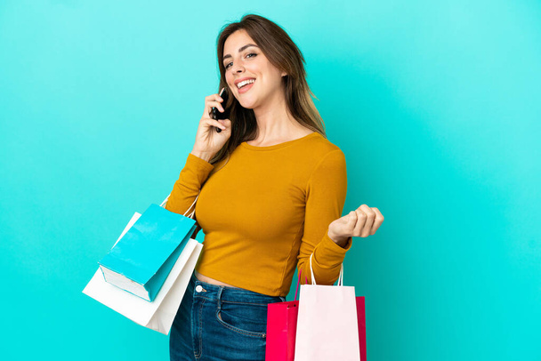 Kaukasische Frau isoliert auf blauem Hintergrund mit Einkaufstaschen und einem Anruf einer Freundin mit ihrem Handy - Foto, Bild