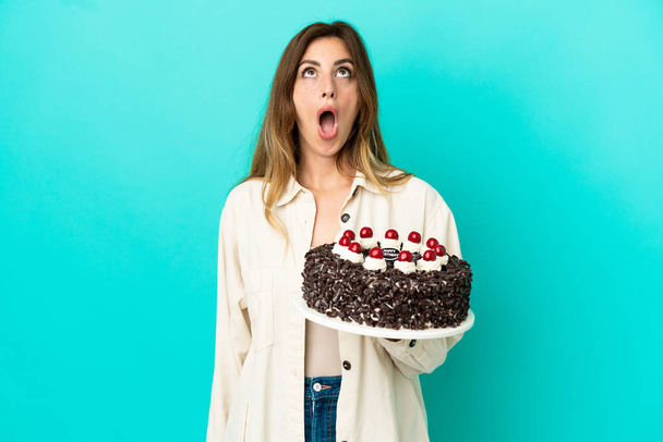 blank vrouw holding verjaardag taart geïsoleerd op blauwe achtergrond op zoek omhoog en met verbaasd expressie - Foto, afbeelding