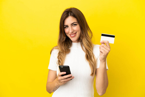 Kaukasierin isoliert auf gelbem Hintergrund kauft mit dem Handy mit Kreditkarte - Foto, Bild