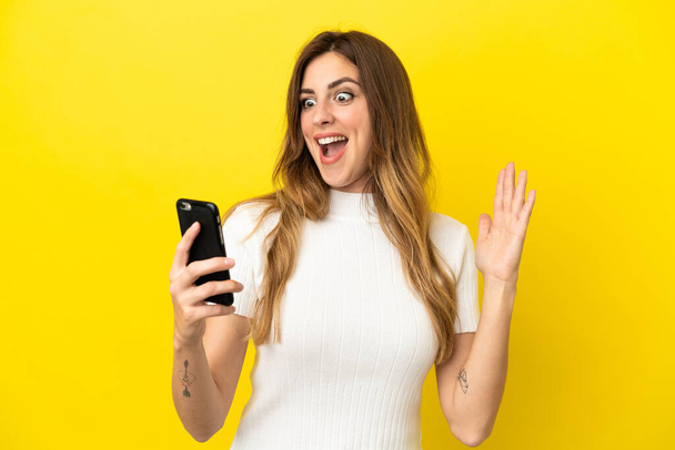 Kaukasische Frau isoliert auf gelbem Hintergrund und schaut in die Kamera, während sie das Handy mit überraschtem Ausdruck benutzt - Foto, Bild