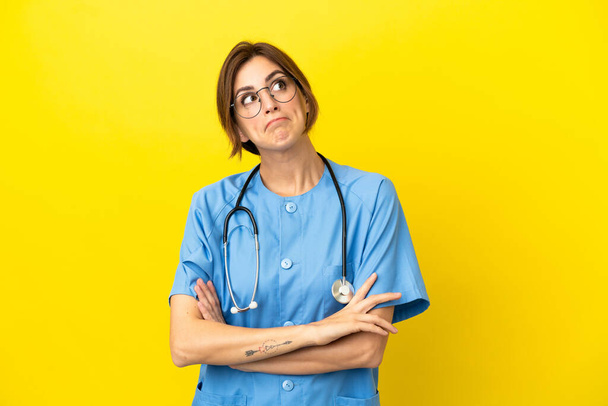 Χειρουργός γιατρός γυναίκα απομονωμένη σε κίτρινο φόντο κάνοντας χειρονομία αμφιβολίες, ενώ άρση των ώμων - Φωτογραφία, εικόνα