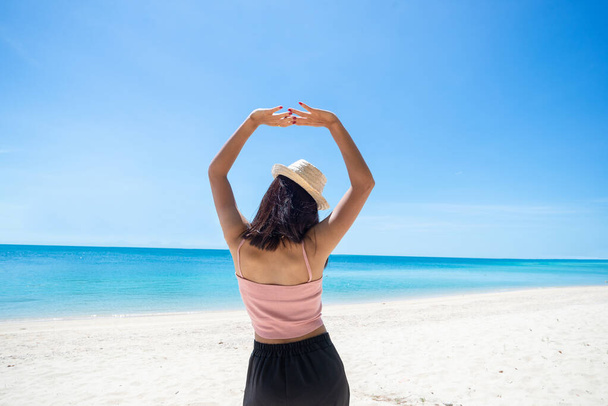 ピンクのタンクの上を身に着けている女性の日焼け肌の背中と空に突き出た腕を持つわらの帽子。海と新鮮な空を見ています。夏の旅行。リラックス、休日と熱帯、快適なコンセプト. - 写真・画像