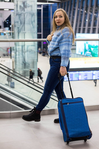 A beautiful stylish woman in casual clothes walks with a blue suitcase through the airport. The concept of tourism and travel - Foto, Imagem