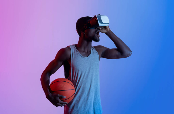 Sanal egzersiz konsepti. Bin yıllık heyecanlı siyah atlet VR kulaklığı takmış elinde neon ışıkla basketbol oynuyor. - Fotoğraf, Görsel