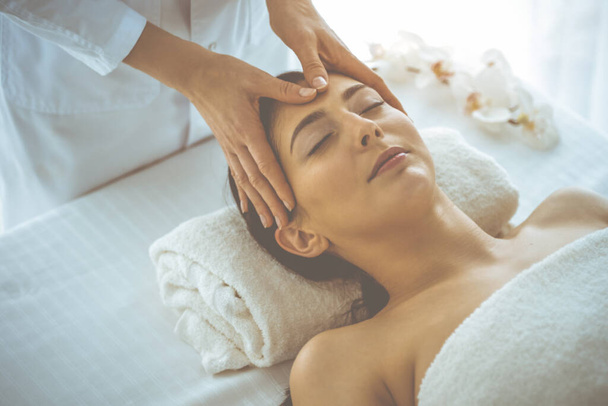 Beautiful brunette woman enjoying facial massage with closed eyes in sunny spa center. Relaxing treatment and cosmetic medicine concepts - Zdjęcie, obraz