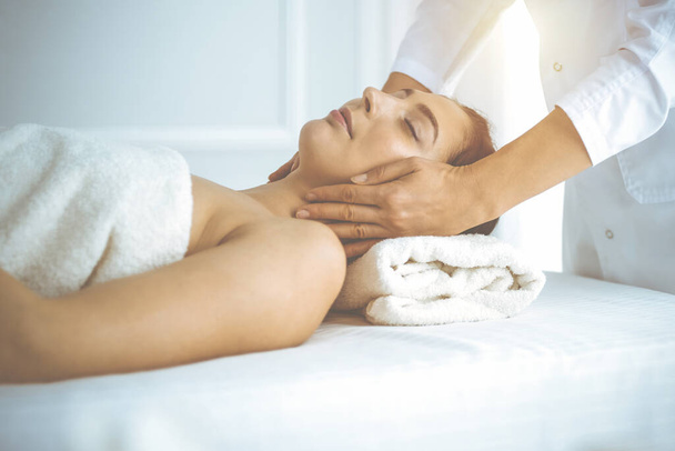 Beautiful brunette woman enjoying facial massage with closed eyes in sunny spa center. Relaxing treatment and cosmetic medicine concepts - Foto, Imagem