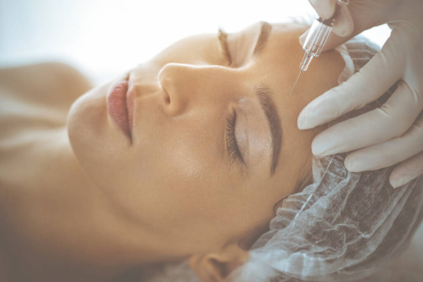 Beautician doing beauty procedure with syringe to face of young brunette woman in sunny clinic. Cosmetic medicine and surgery, beauty injections - Valokuva, kuva