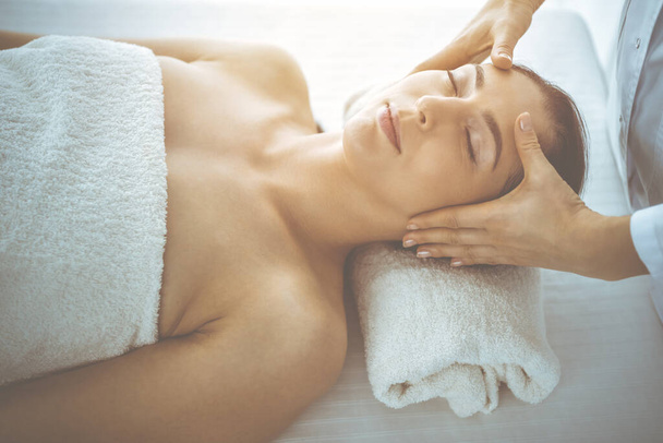 Beautiful brunette woman enjoying facial massage with closed eyes in sunny spa center. Relaxing treatment and cosmetic medicine concepts - Photo, image