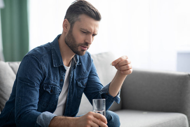 Расстроенный бородатый мужчина со стаканом воды, принимающий лекарства - Фото, изображение