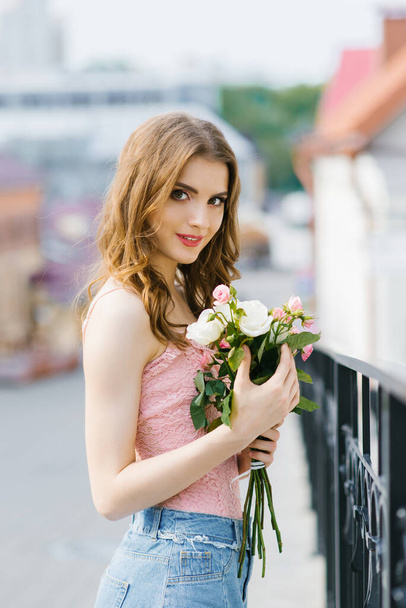 Ein blondes junges hübsches Mädchen in Sommerkleidung steht da und hält nach einem Date zarte weiße und rosa Rosen in den Händen - Foto, Bild