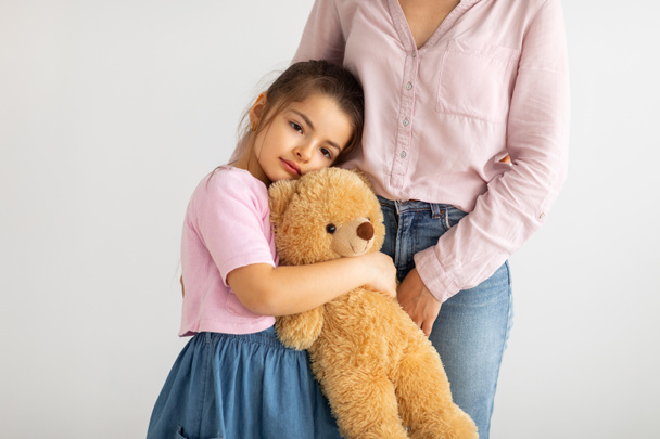 Cute little girl with teddy bear embracing mother and standing together against light wall - Photo, Image